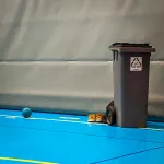 En grå soptunna med återvinningssymbol och ett par skor på ett blått golv inne i en sporthall.