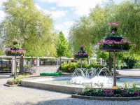 En springbrunn sprutar vatten omgiven av blomsterpelare med rosa geranium i en solig stadspark med pilträd och kullerstensplattor.
